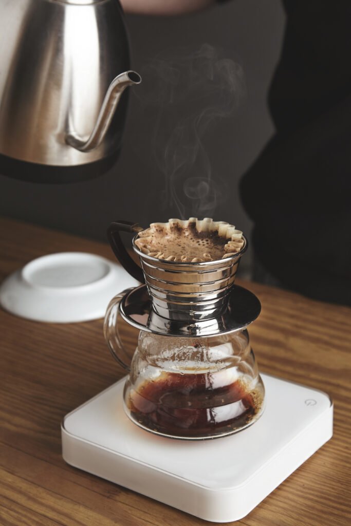 Barista in black sweatshot prepares filtered coffee/silver teapot to beautiful transparent chrome drip coffee maker on white simple weights. Everything on thick wooden table in cafe shop. Steam