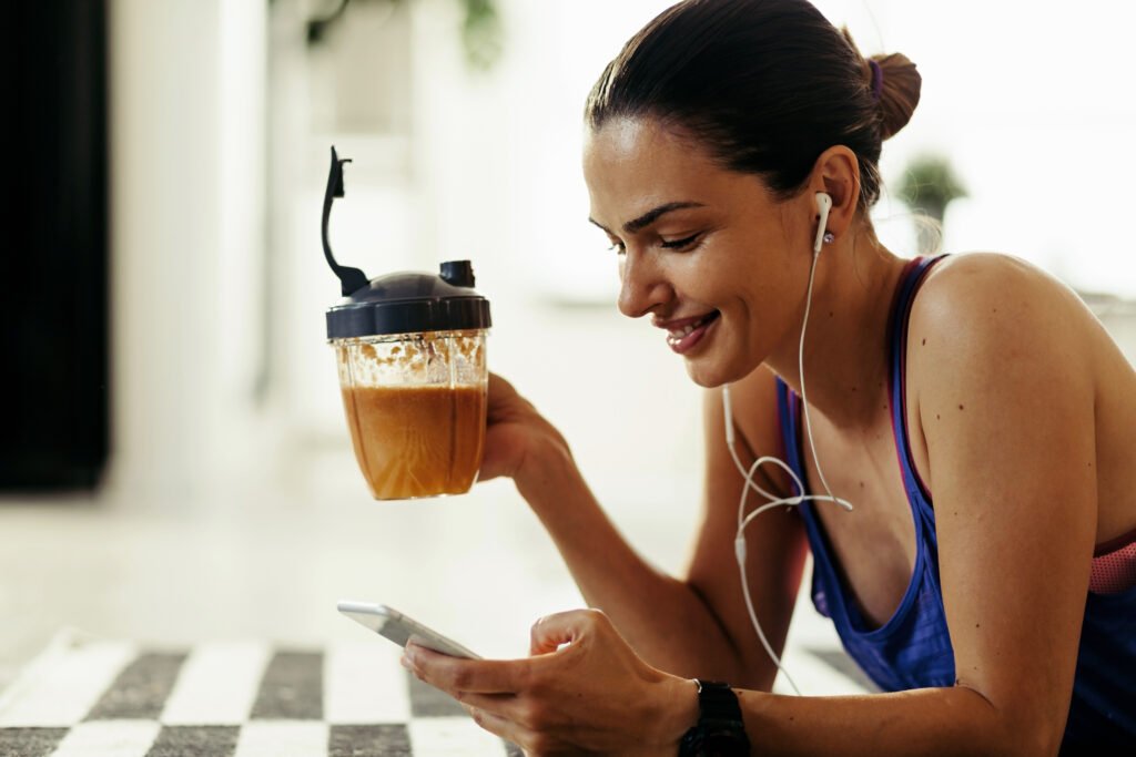 Happy sportswoman text messaging on smart phone while lying down on her benny on the floor and drinking coffee - best times to drink coffee