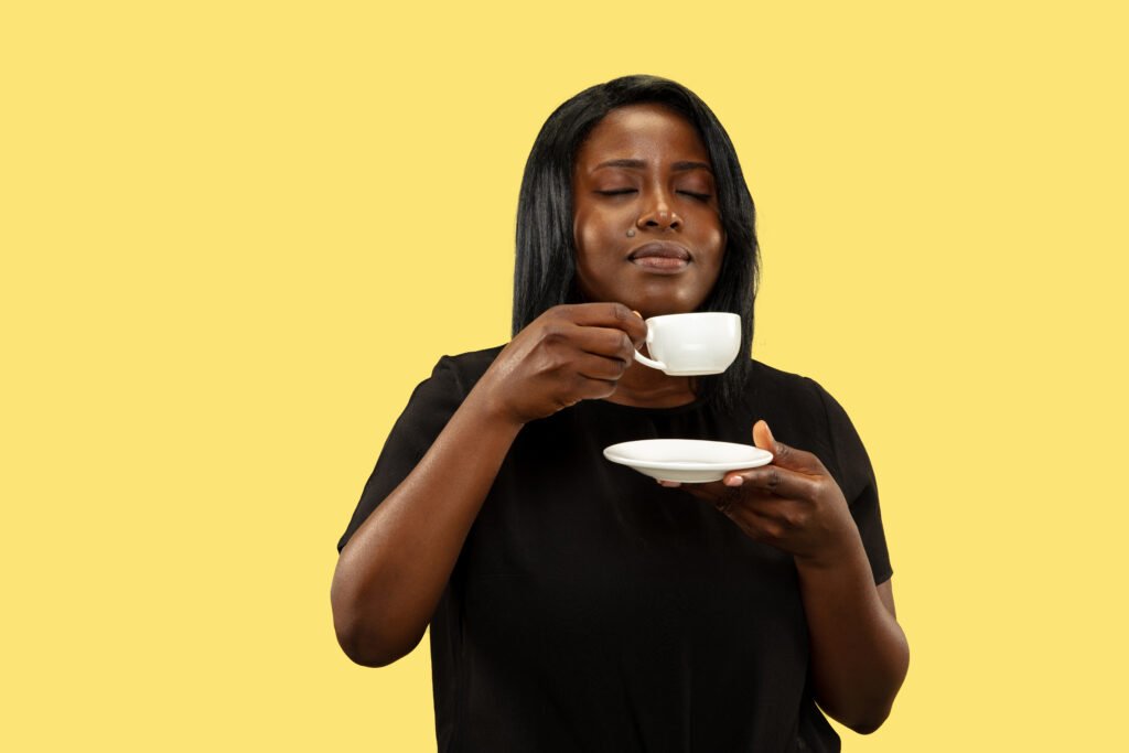 Young African-American woman isolated on yellow studio background, facial expression. Beautiful female half-length portrait. Concept of human emotions, facial expression. Enjoying coffee consumption.