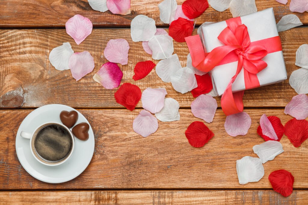  Valentines Day gift in white box amidst white and red petals and a cup of coffee in white mug on white saucer on wooden table - Perfect Valentine’s Gifts for Coffee Lovers 